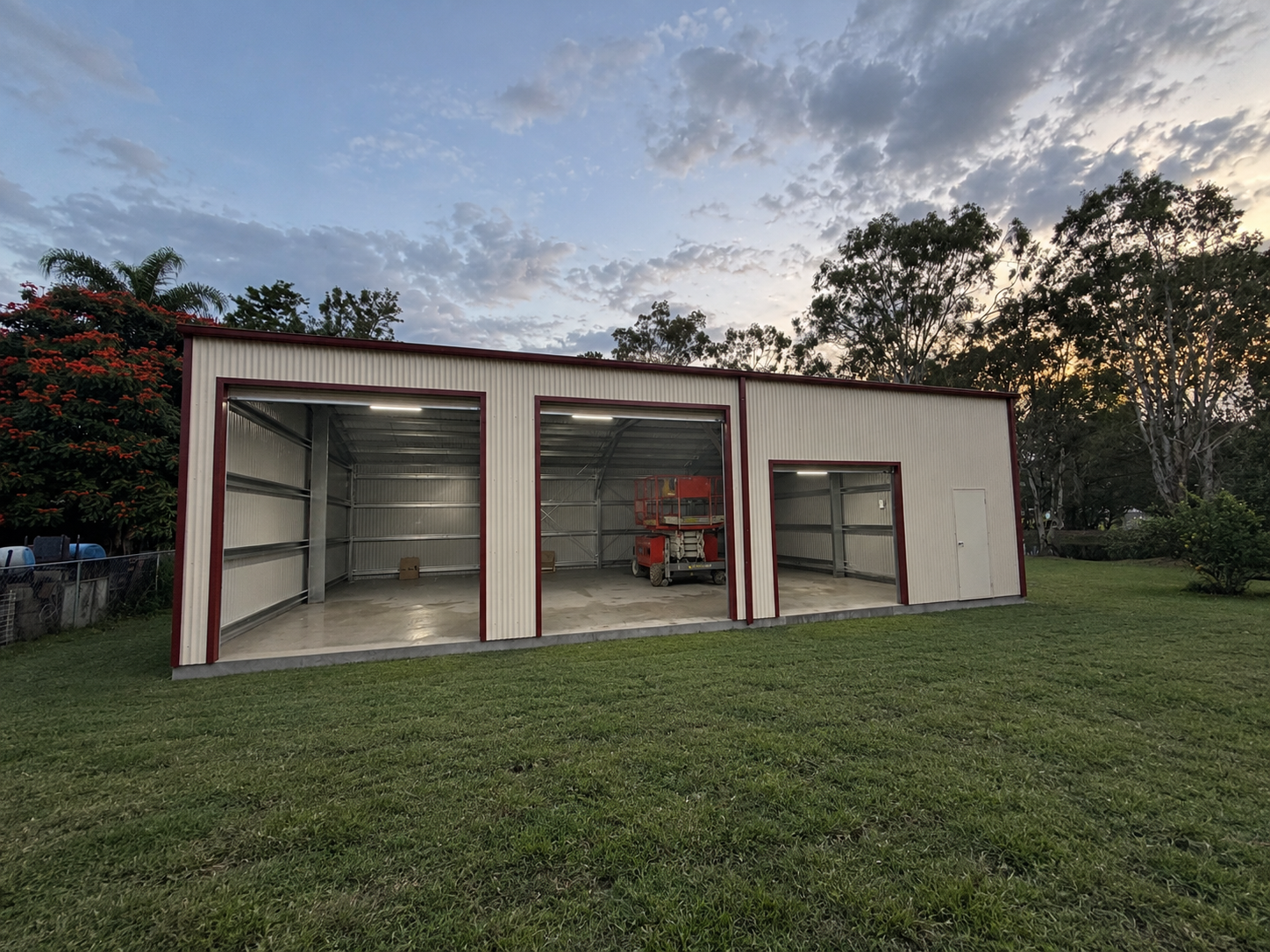 Side view of a three-bay Colorbond workshop shed with open roller doors at dusk, Tamborine